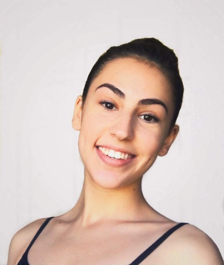 Image Description: Camille is in front of a light, plain backdrop, smiling at the camera. Her dark brown hair is pulled back in a bun and she is wearing a top/leotard with thin black straps.