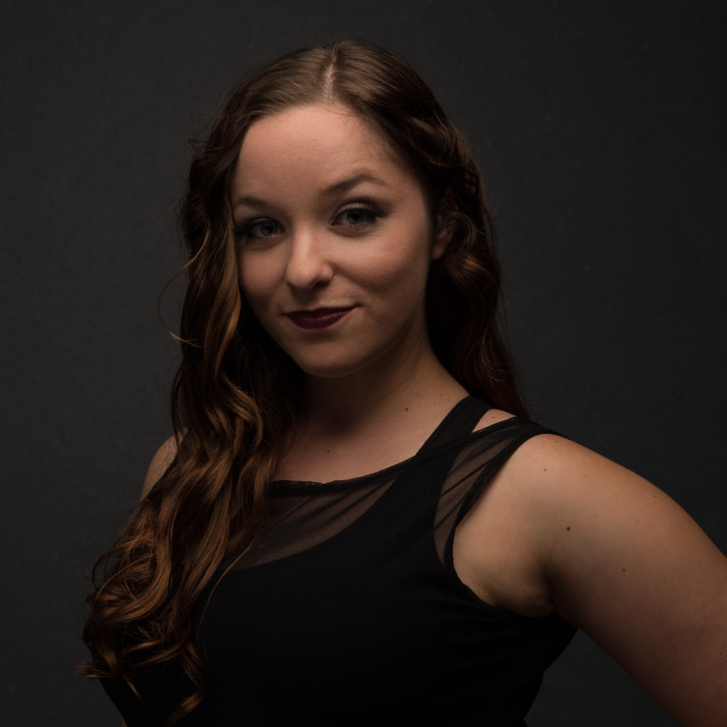 Image Description: A headshot of Arielle against a black backdrop. She has long, curled brown hair that is parted in the middle. She has a slight smile and wears a black mesh tank top with a black athletic shirt underneath.