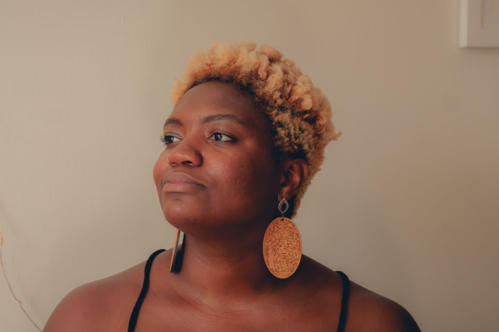 Photo credit: Chidozie Ekwensi. Image description: Photo of a dark skinned Black woman staring towards the light, with her head tilted upwards at an angle. She has short curly blonde hair and wears large wooden earrings