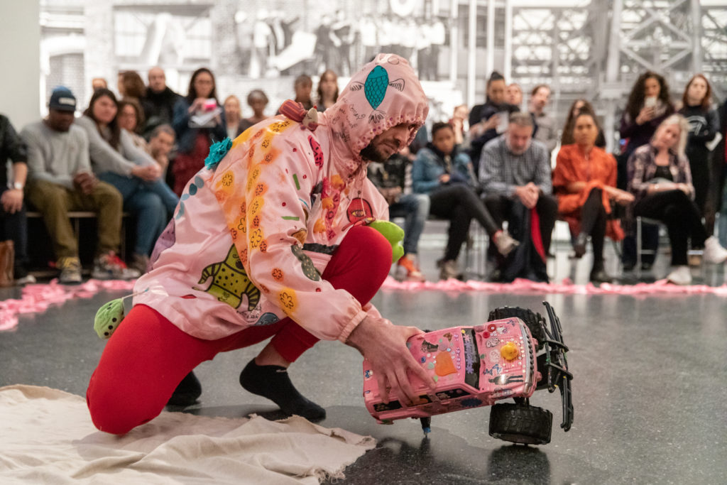 Image Description: Christopher is crouching and holding a pink-painted remote control car. His costume is a pink hoodie and red pajamas. Behind is the audience paying close attention to his performance.
Photo credit: Walter Wlodarczyk, courtesy of the Immigrant Artist Biennale.