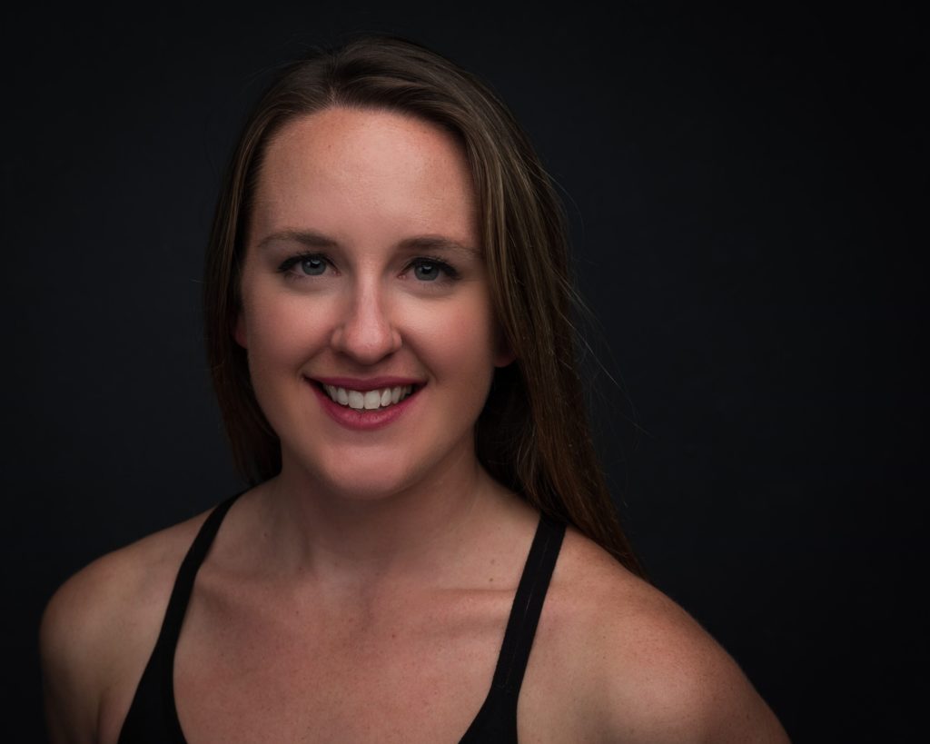 Image Description: Jessica smiles at the camera against a black backdrop. Her long hair is down and she wears a black athletic tank top.
