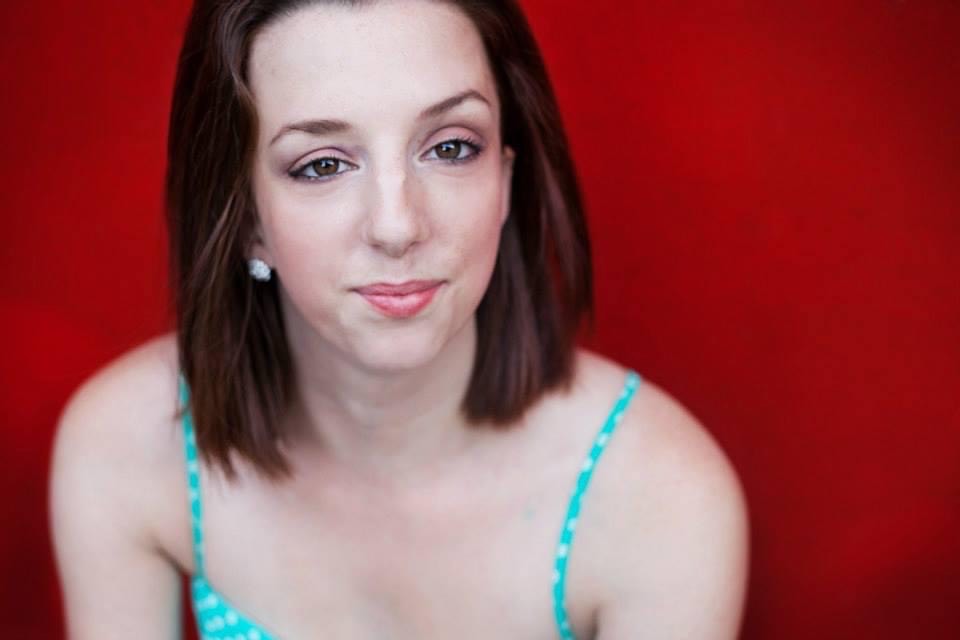 Louisa smiles for the camera; she has straight auburn hair, pale skin, brown eyes and arched brown eyebrows. She is wearing a turquoise blue camisole and poses against a red background.