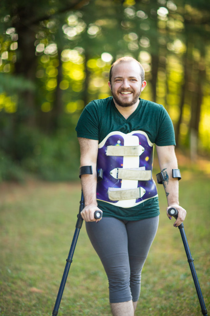 Image description: Toby, a short, broad-shouldered white person with a buzzed head and short dark beard, stands casually leaning into their forearm crutches, a galaxy-print back brace over their leggings and t-shirt. They are outdoors, with a backdrop of soft-focus trees in summer greens and golds. They are smiling directly toward camera.