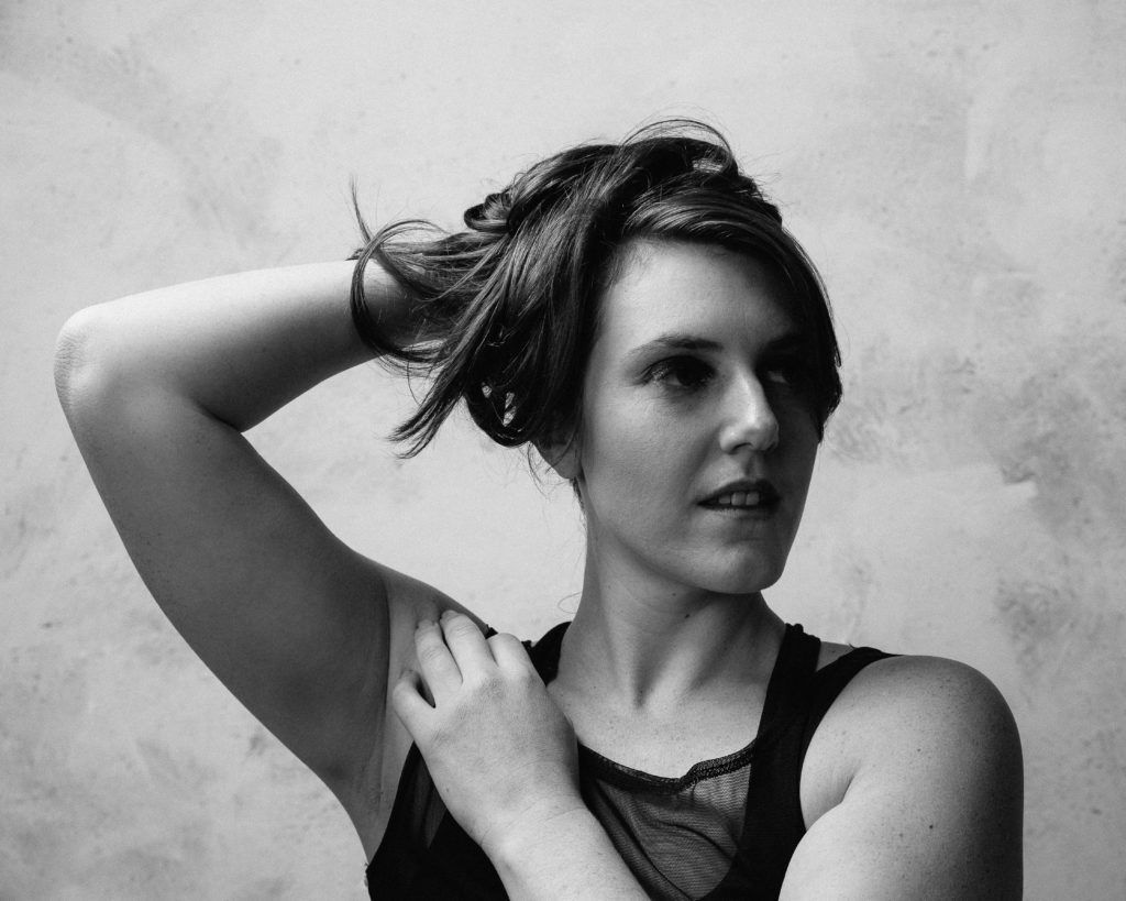Image Description: Black and white headshot of Heather with one arm across her body resting on shoulder, the other holding her long brown hair behind her head. Her gaze looks off to the side of the camera. Heather wears a black mesh tank top.