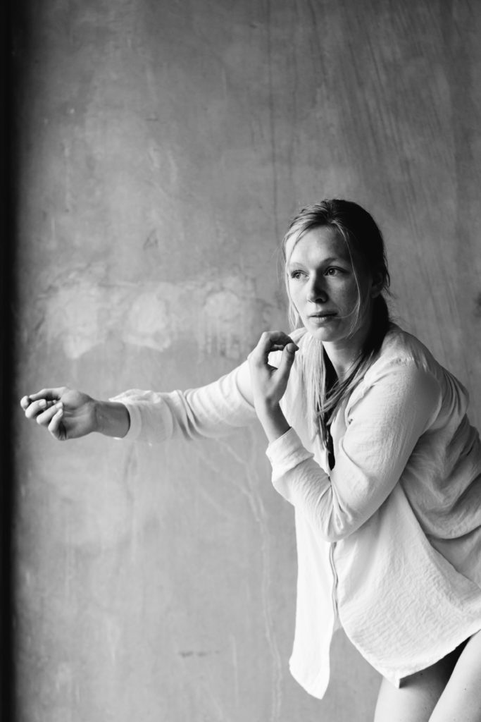 Image description: A black and white photograph of Mayke, her light colored hair is back and she wears a baggy light colored button down top with 3/4 length sleeves. She reaches one arm forward with a soft fist, while the other hand is lifted a few inches from her chin. Her gaze extends beyond her hands with a serious expression.