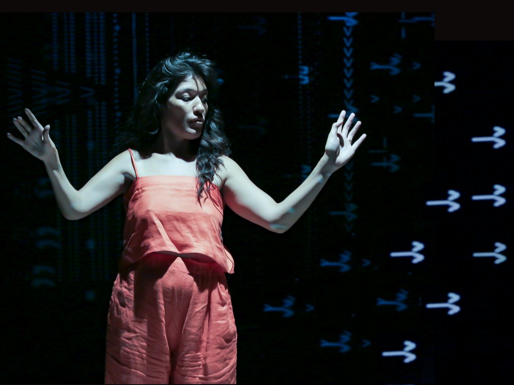 Pelenakeke, a brown skinned woman with long wavy brown hair, wears an orange top and pants, looks to the side with both arms lifted, creating a W shape.