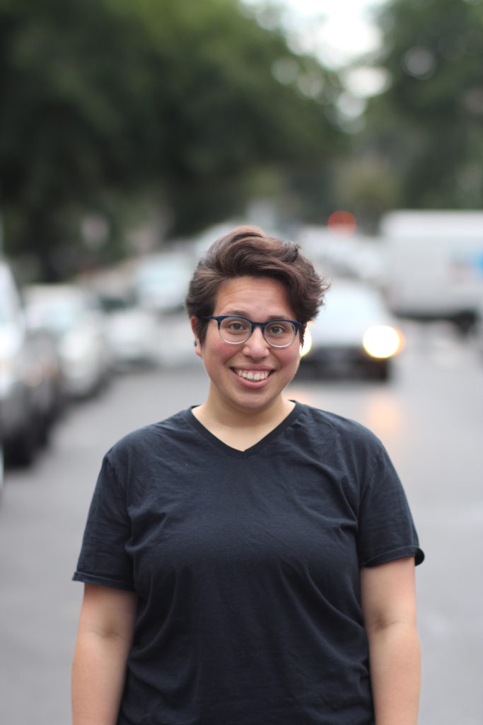 M, a large-bodied, white, Latinx person, stands in a relaxed posture smiling directly at the camera. They are wearing a wrinkled black t-shirt and two-toned blue and clear glasses. They are standing in the middle of a tree-lined road on a cloudy day with cars parked on either side of them. M is sharply in focus while the objects behind them are blurry. In this stolen shot, a car is approaching from directly behind them. Photo credit: Travis Jones 