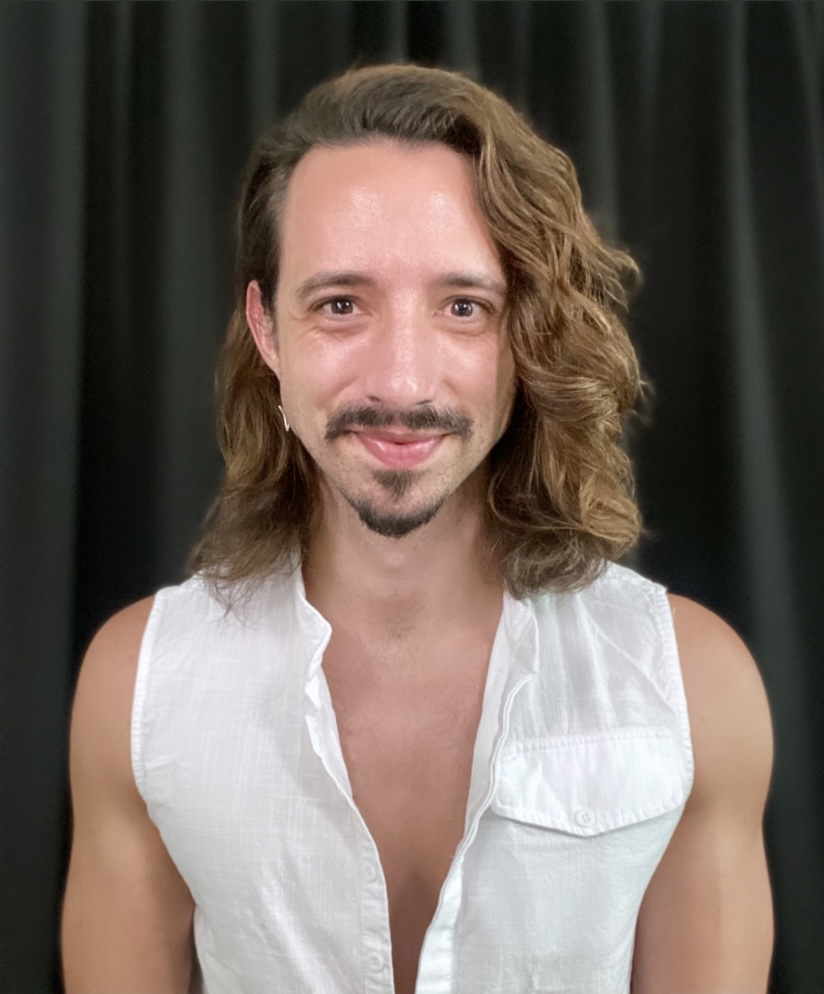 Image description: A headshot of Cameron with a closed mouth smile and shoulder length hair. He wears a white v neck tank top.