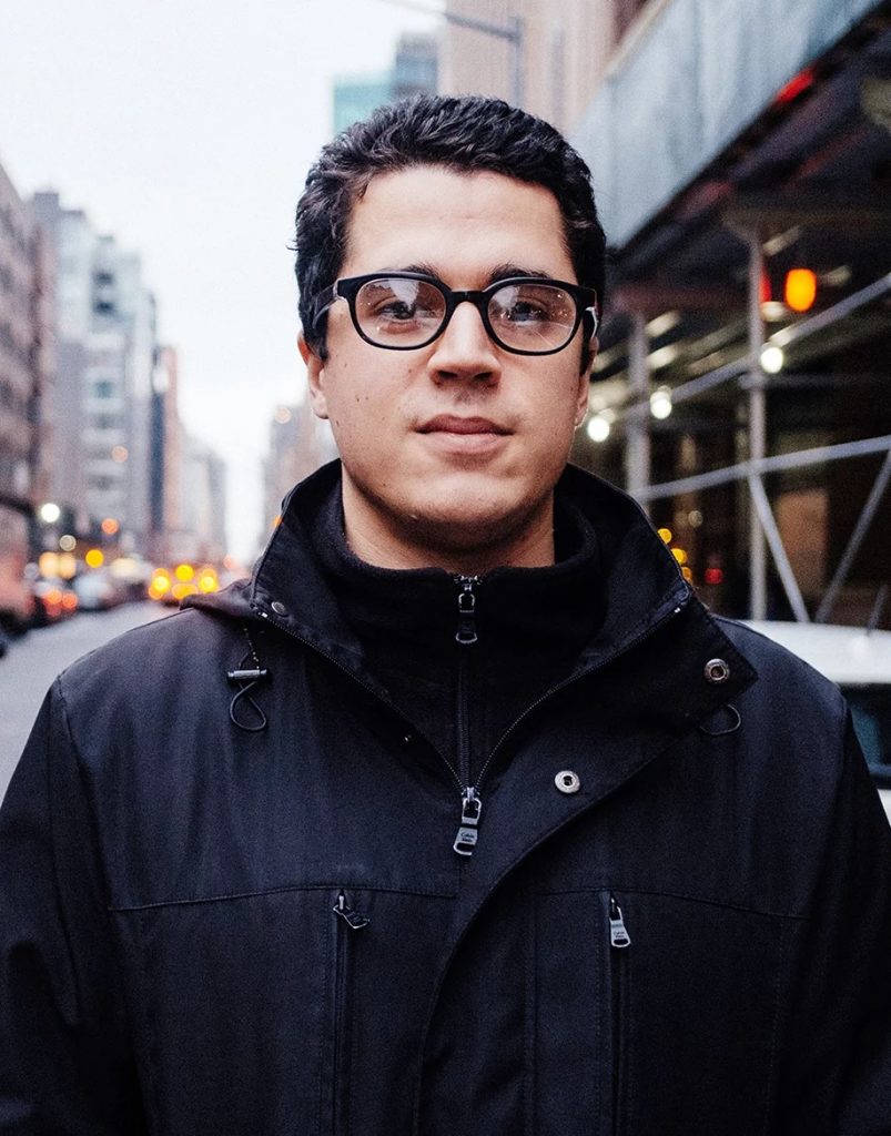 Headshot of Ryan standing on a busy city street. Ryan is looking straight at the camera and wears a black jacket and black rimmed glasses.