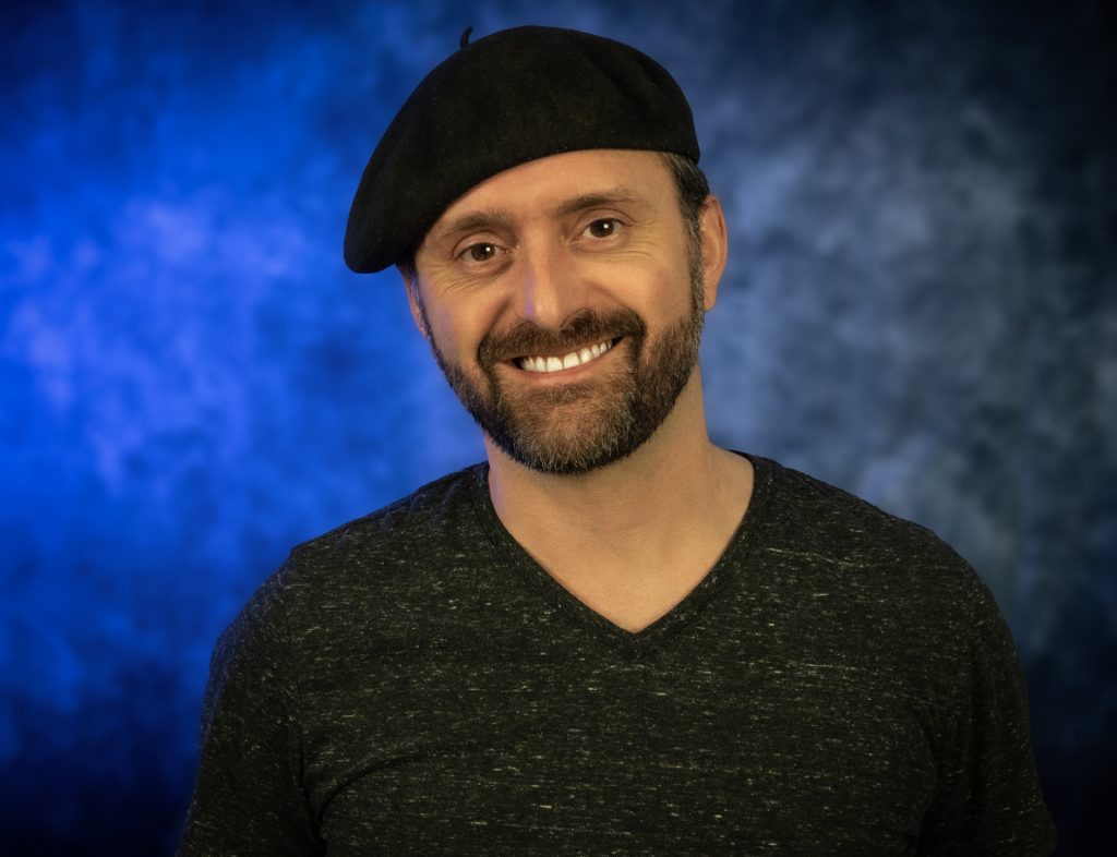 Ian is in front of a blue background, smiles at the camera, wearing a grey v-neck and black bureau hat.