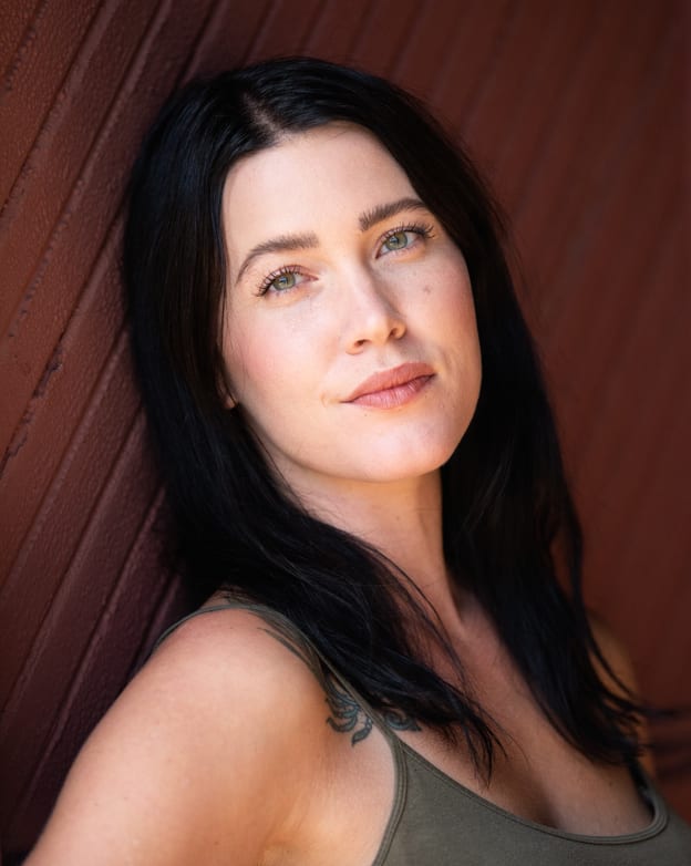 Headshot of Abigail, leaning on a deep red wall, her dark hair is down and she looks to the camera. 